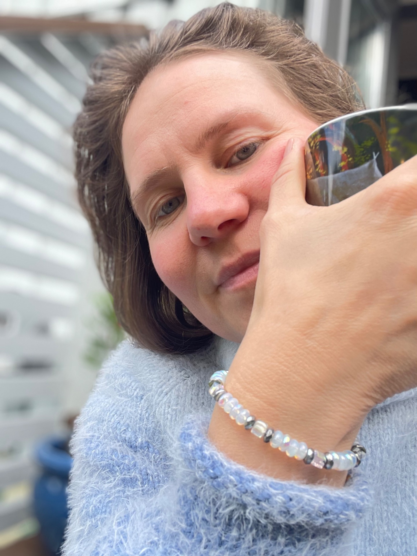 Nærbilde av kvinne med hånd ved kinnet, iført blå genser og armbånd, sollys og uskarp bakgrunn.