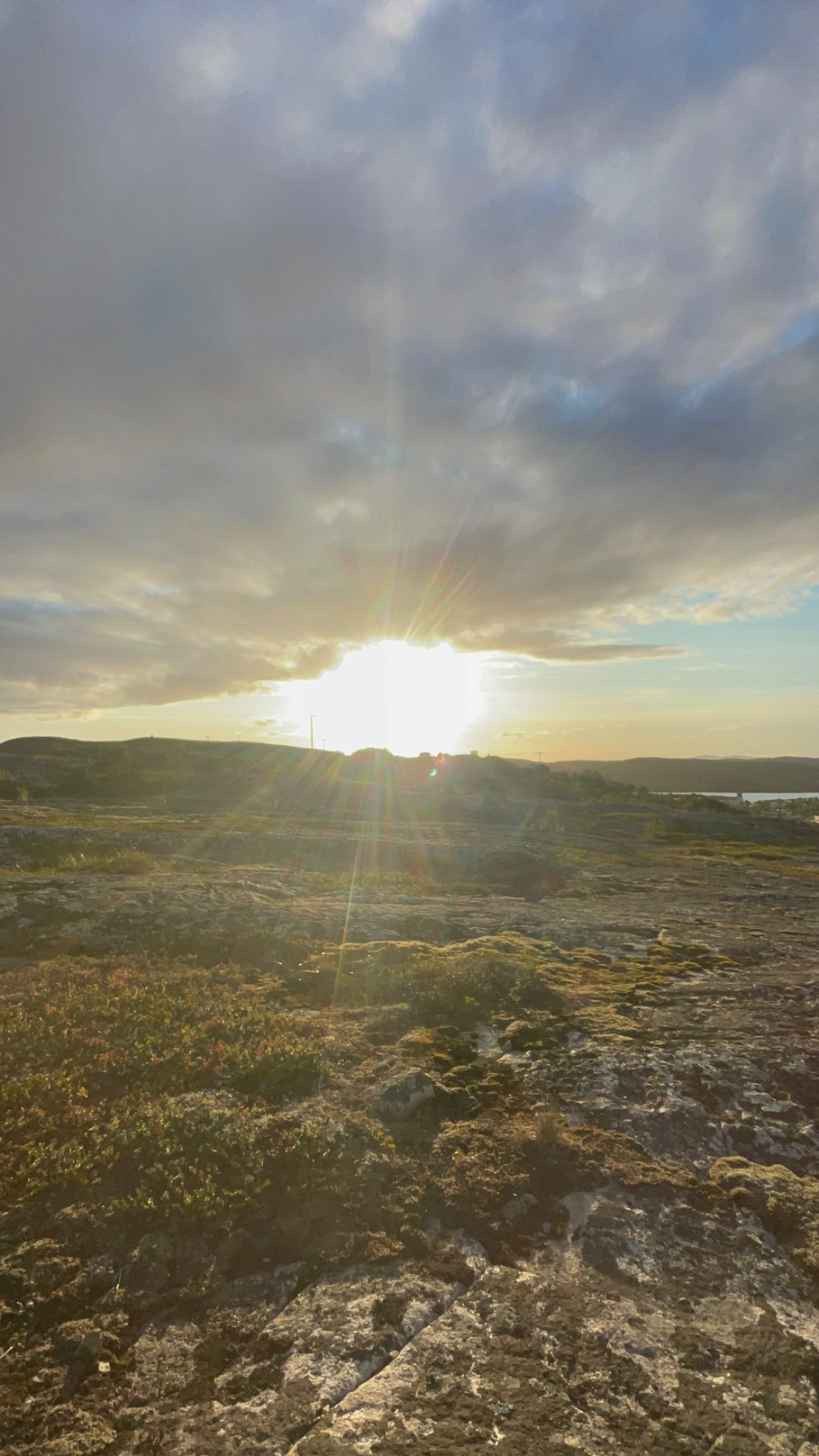 Solformørkelse over kystlandskap med bølgende åser, steinete forgrunn og stråler gjennom skyene.